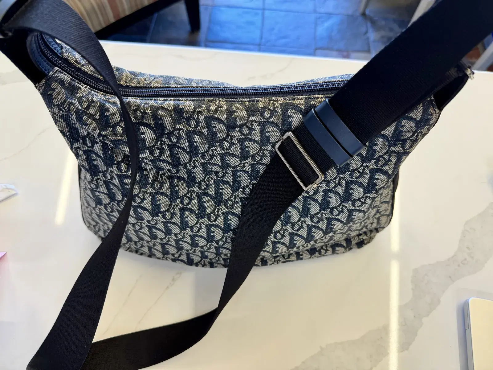 Back of the cleaned Dior Trotter messenger bag with its black webbing strap and navy leather tabs, the jacquard along the zipper clean and even